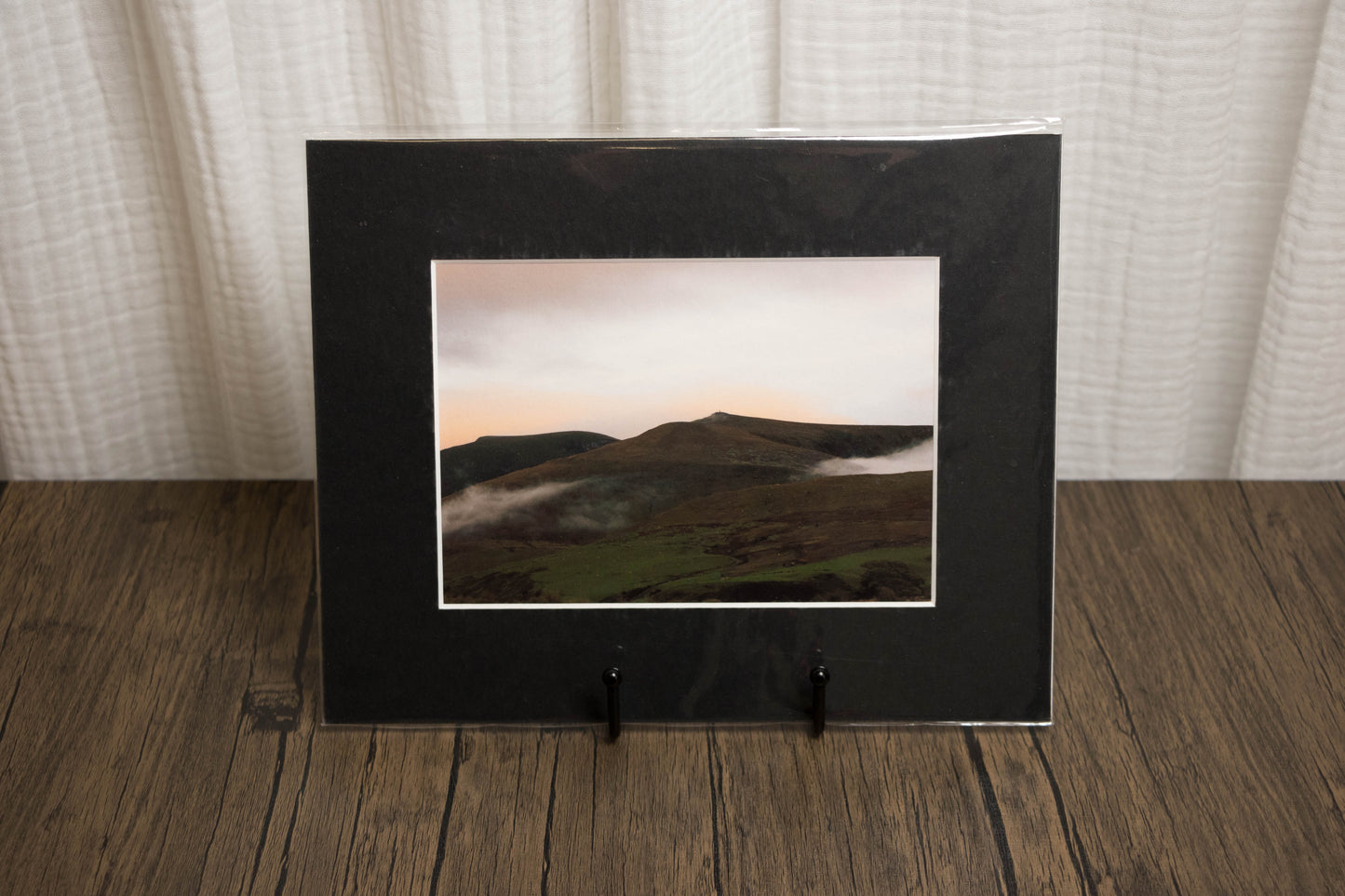 Framed landscape photo on a wooden surface with a white curtain background