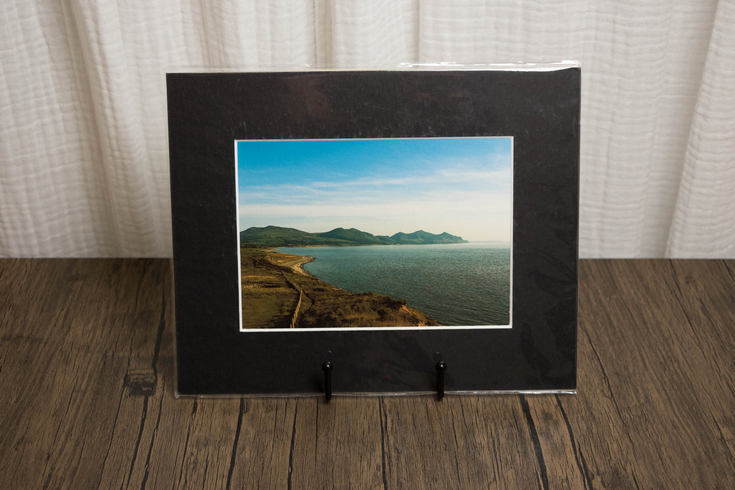 Framed photograph of a coastal landscape on a wooden surface with a white curtain background.