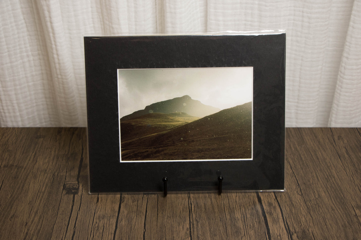 Black framed photograph of a mountain landscape on a wooden surface with a white curtain background.