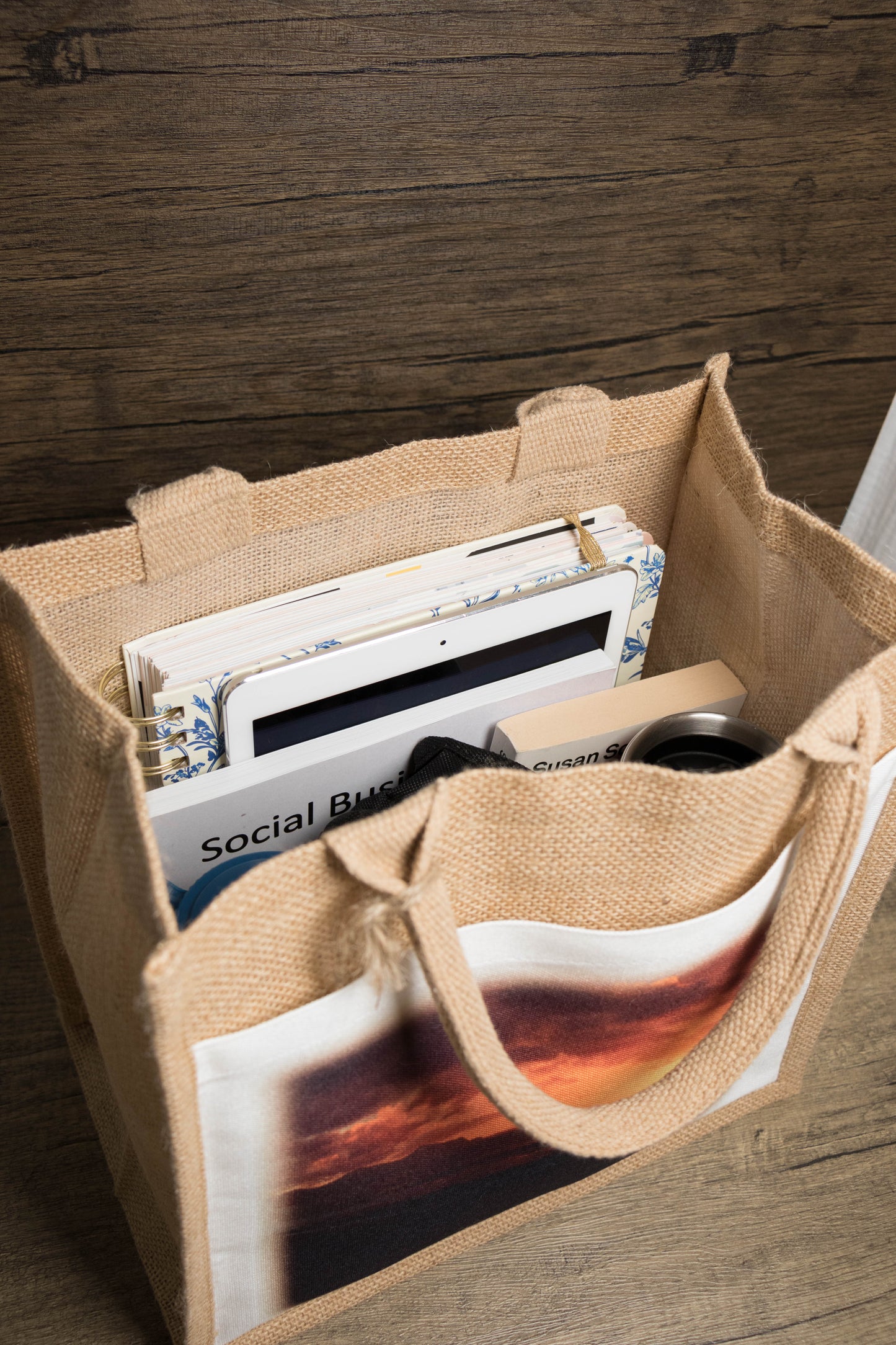 jute bag with printed design on the pocket filled with waterbottle, pencilcase, drink flask, three books and a tablet.