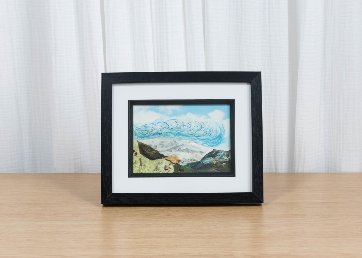 Framed artwork on a wooden surface with a white curtain background