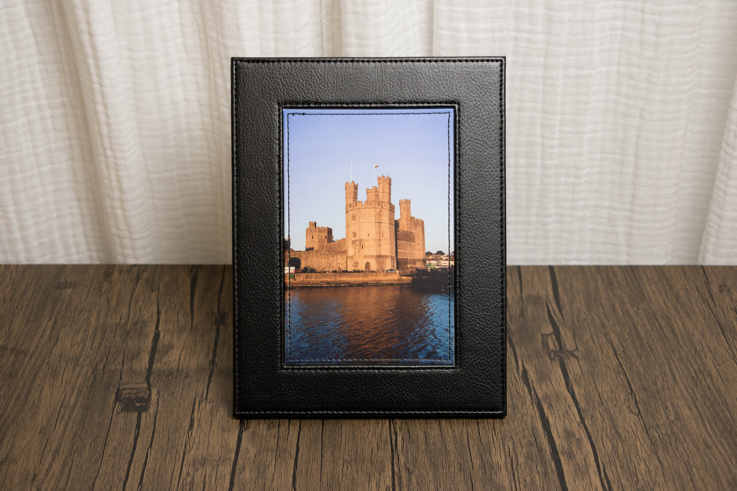 Framed picture of Caernarfon castle on a wooden surface with a white curtain background