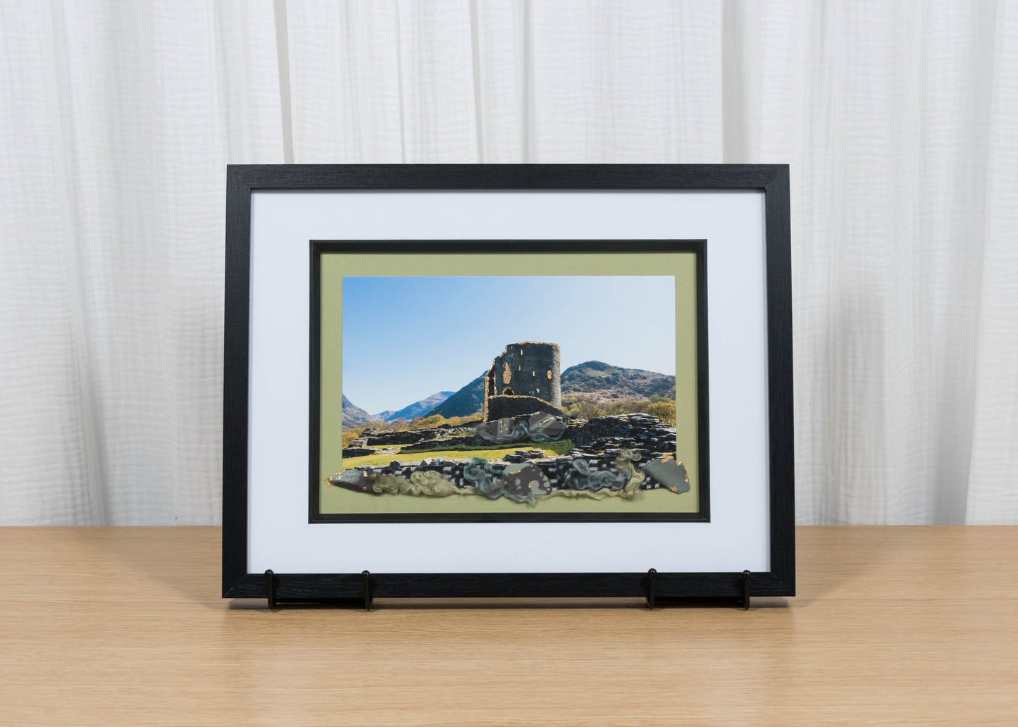Framed artwork of a landscape with a castle on a wooden surface and white curtain background