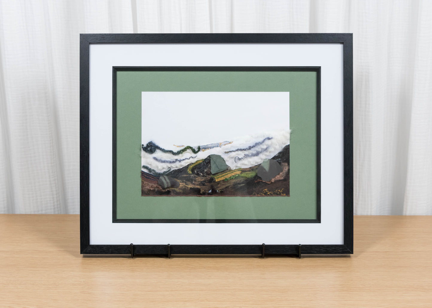 Framed artwork of a mountain landscape on a wooden table with a white curtain background