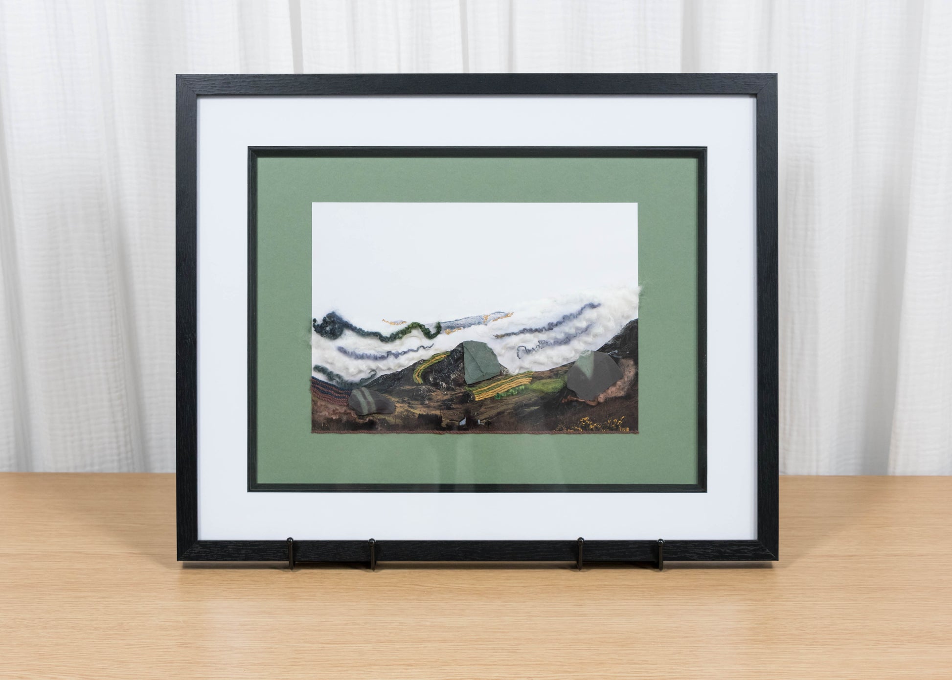 Framed artwork of a mountain landscape on a wooden table with a white curtain background