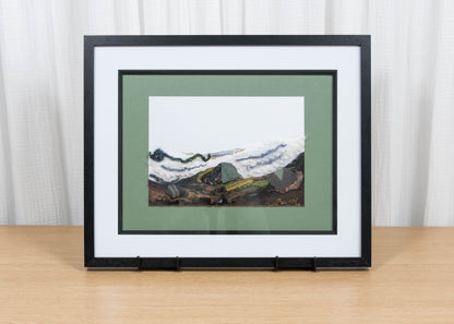 Framed artwork of a mountain landscape on a wooden table with a white curtain background