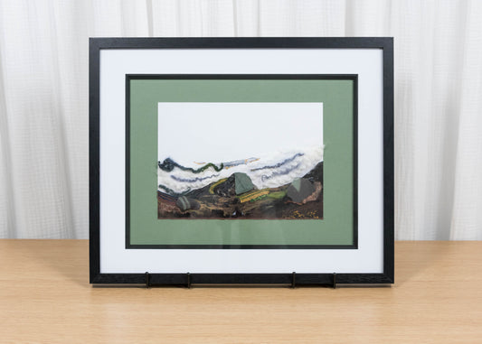 Framed artwork of a mountain landscape on a wooden table with a white curtain background
