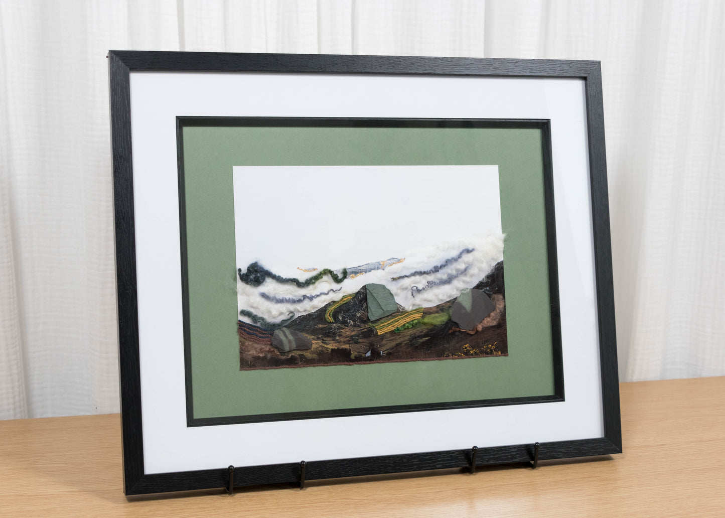 Framed artwork of a mountain landscape on a wooden table with a white curtain background