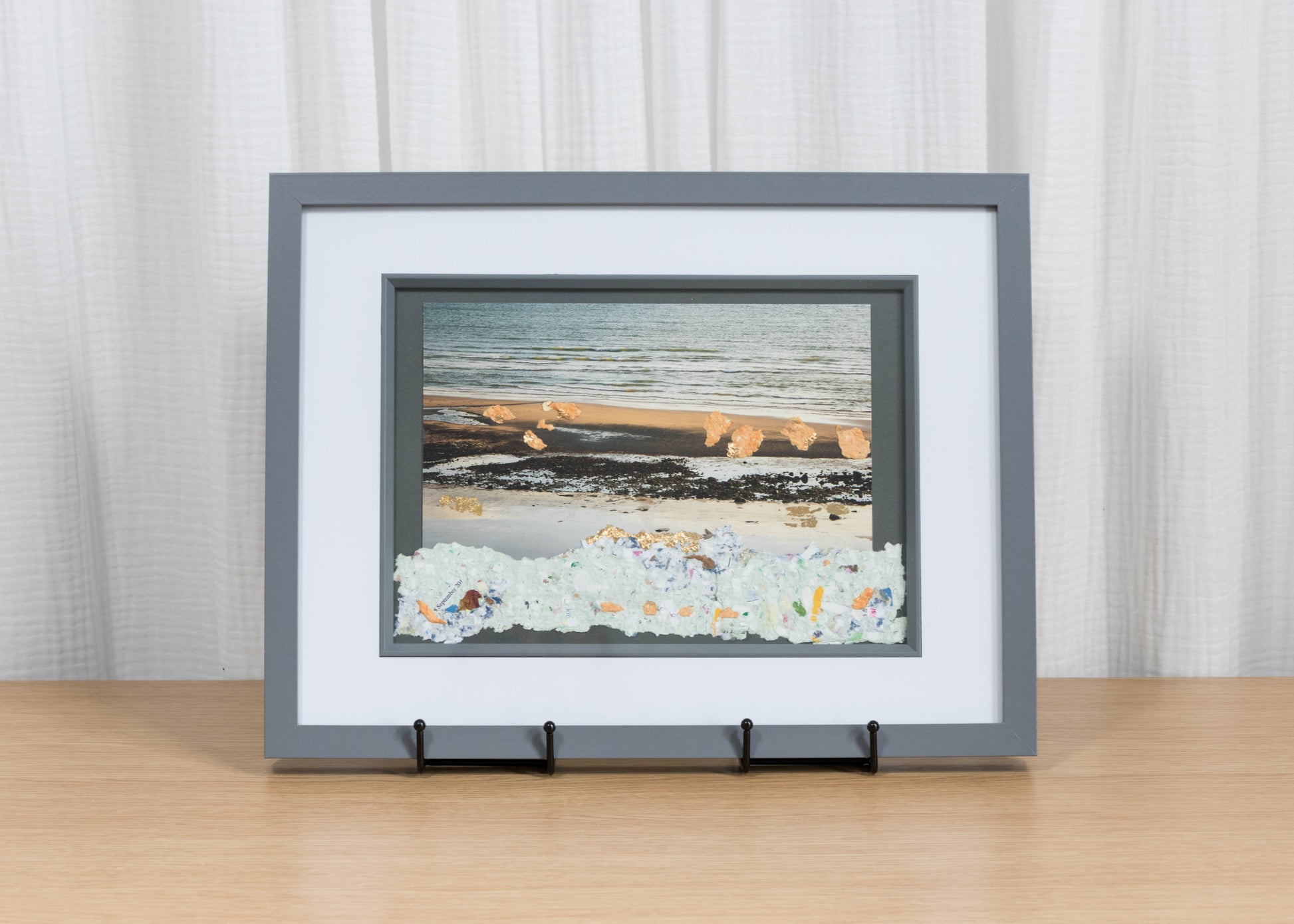 Framed artwork of a beach scene on a wooden surface with a white curtain background