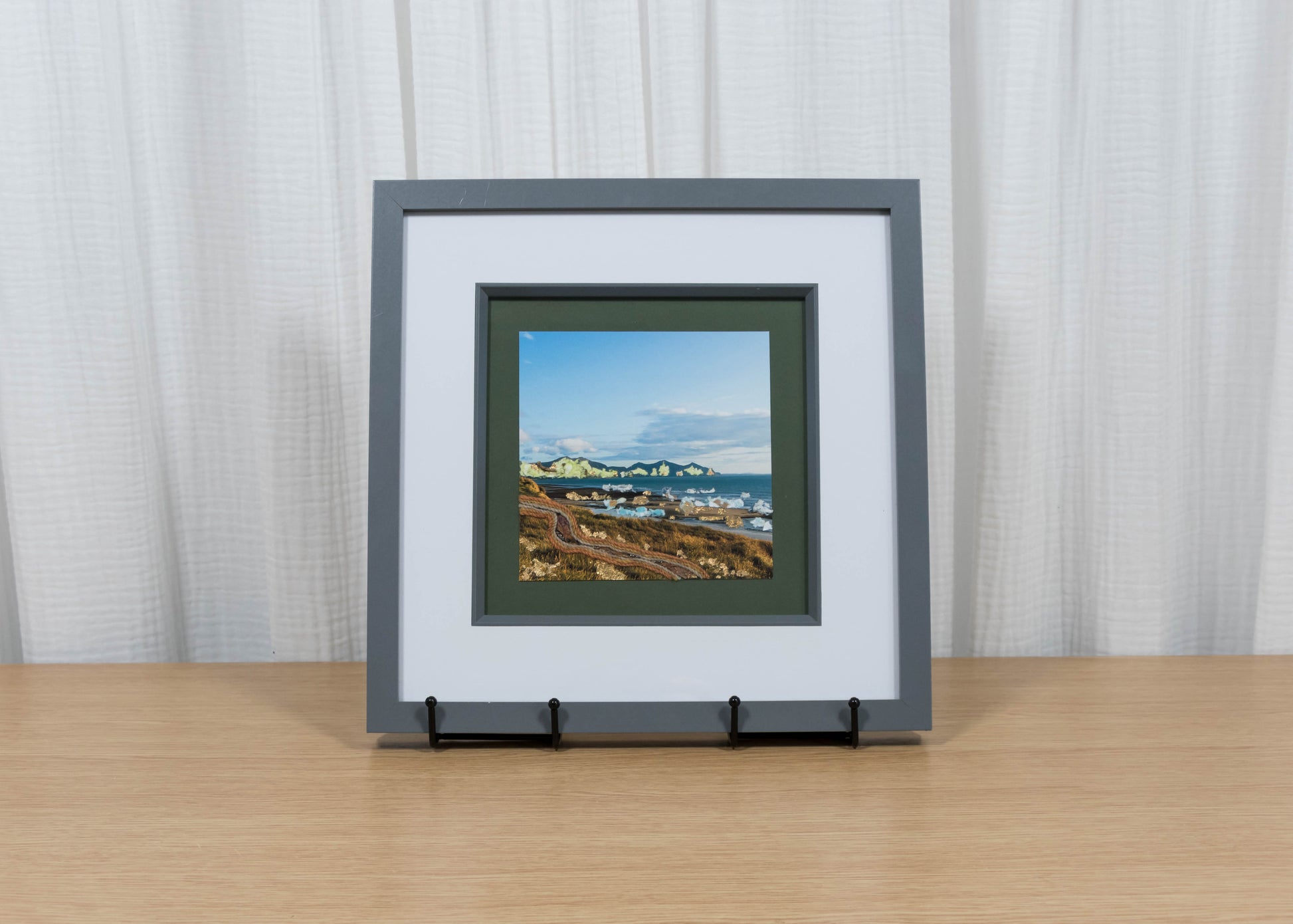 Framed artwork of a coastal scene on a wooden table with a white curtain background