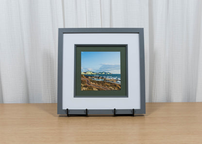Framed artwork of a coastal scene on a wooden table with a white curtain background