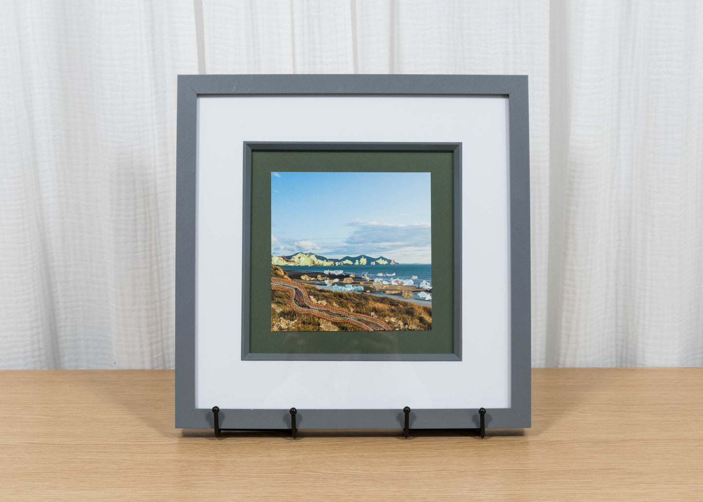 Framed artwork of a coastal scene on a wooden surface with a white curtain background