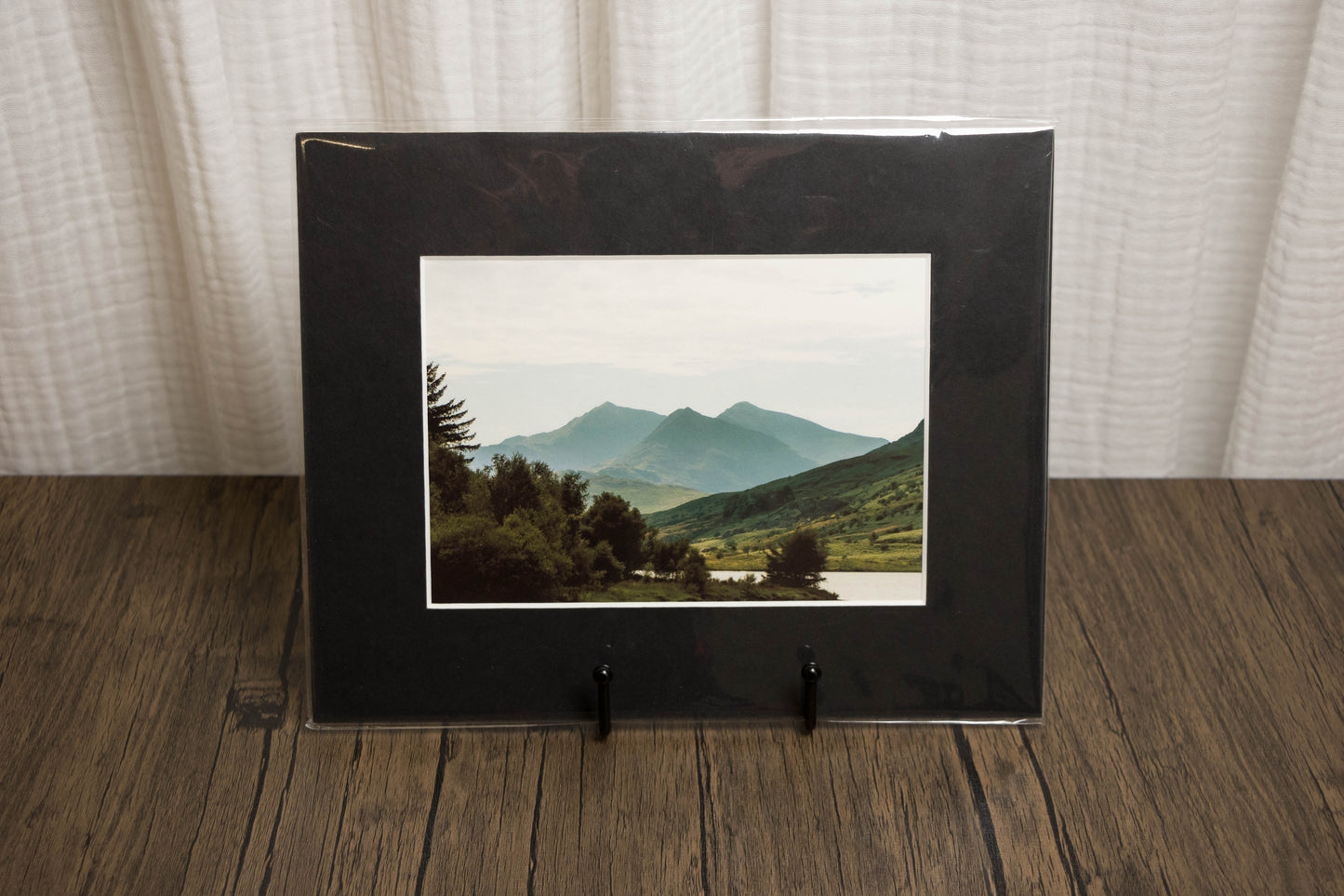 Black framed photograph of a mountain landscape on a wooden surface with a white curtain background.