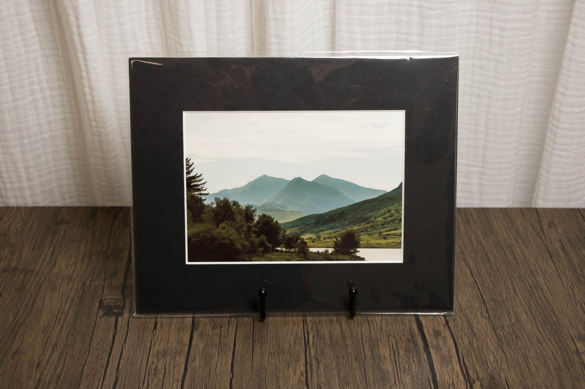 Black framed photograph of a mountain landscape on a wooden surface with a white curtain background.