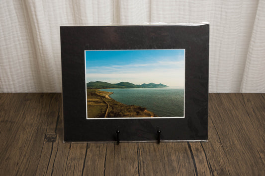 Framed photograph of a coastal landscape on a wooden surface with a white curtain background.