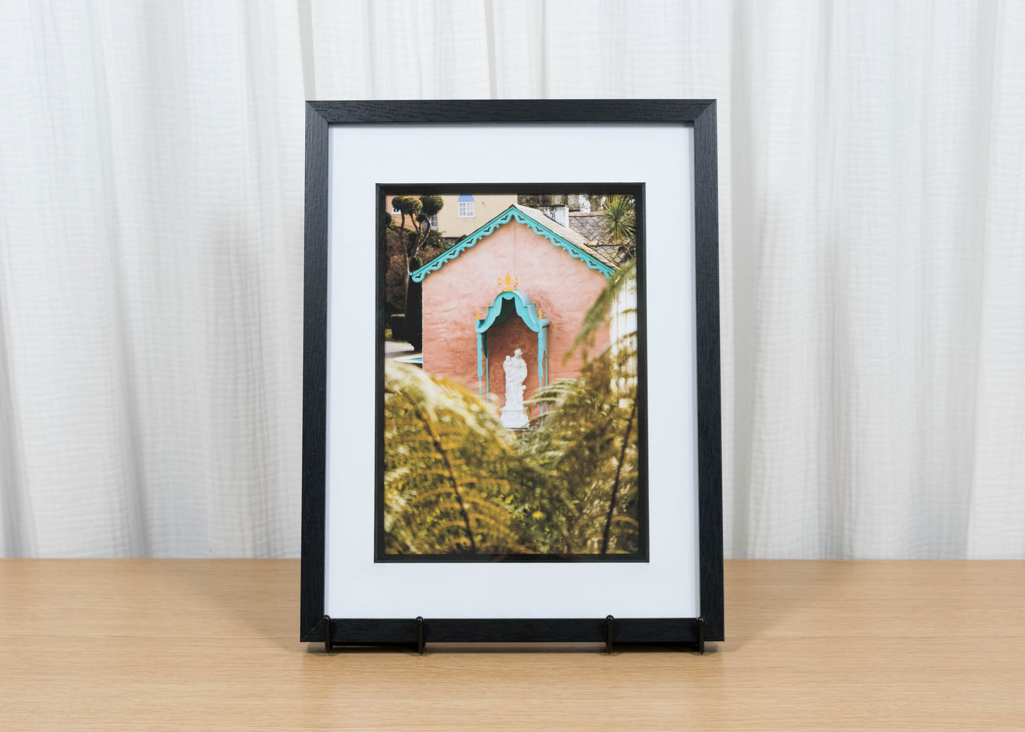 Framed photograph of a pink building with a statue on a wooden surface and white curtain background
