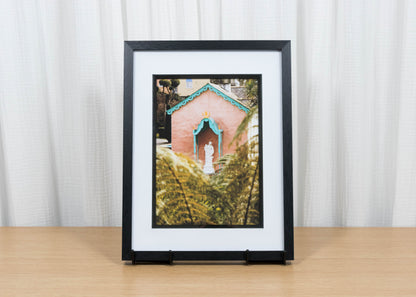 Framed photograph of a pink building with a statue on a wooden surface and white curtain background
