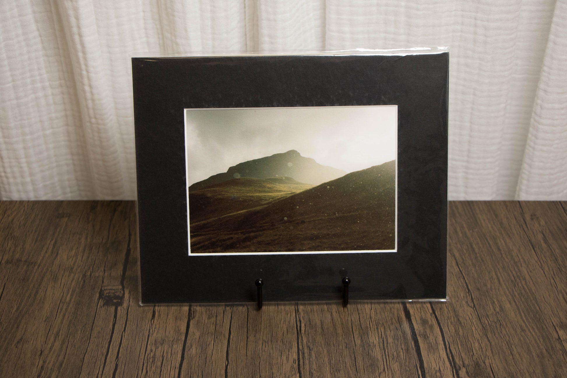 Black framed photograph of a mountain landscape on a wooden surface with a white curtain background.