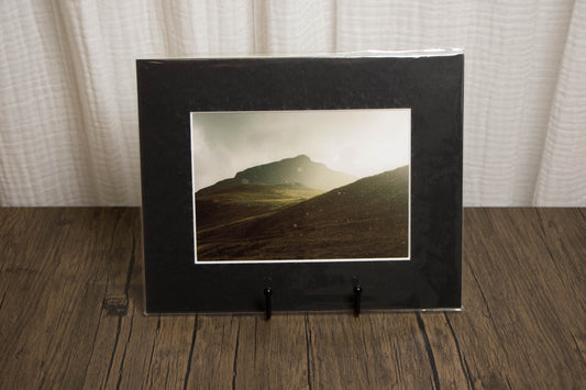 Black framed photograph of a mountain landscape on a wooden surface with a white curtain background.