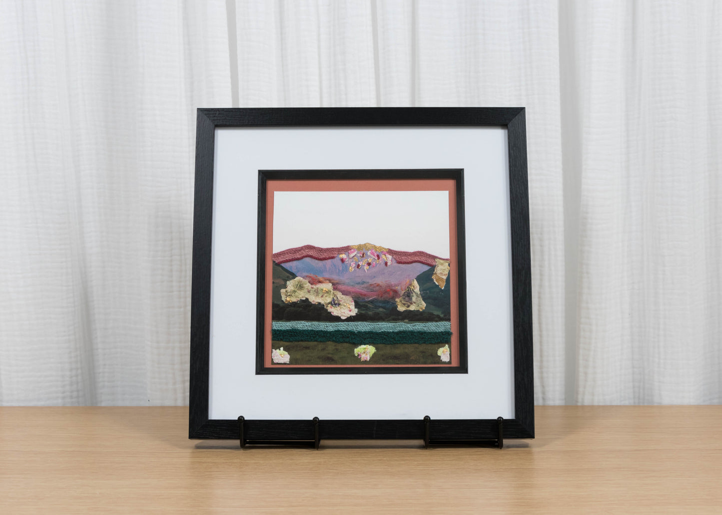 Framed artwork of a mountain scene on a wooden table with a white curtain background